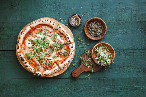 Vegetarian pizza with tomato sauce, sun-dried tomatoes, vegan cheese, microgreens,  seeds and nuts. Flat lay top-down composition on dark green background.