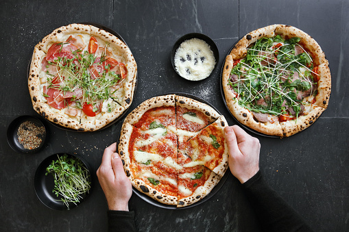 Man eating Italian steak pizza with tomato, arugula, microgreen and pesto sauce. Pizza Margherita. Ham and cheese pizza. Flat lay top-down composition on dark background.