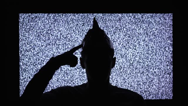Silhouette of a man wearing tin foil hat in a front of TV