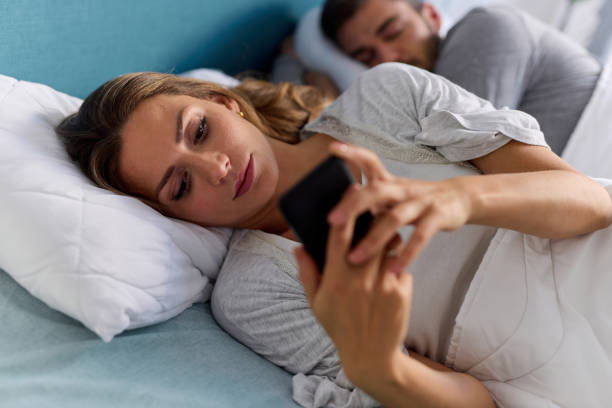 istock Young woman using mobile phone and checking messages in bed while her husband is asleep 1366575352