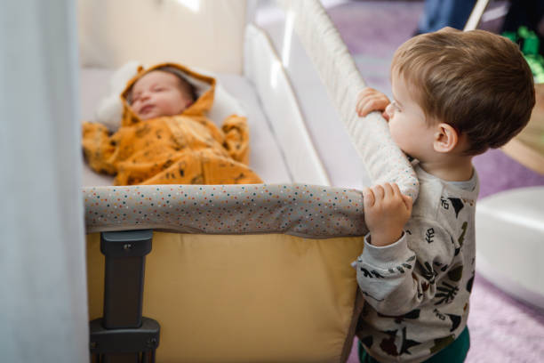 A toddler curiously looking at his newborn brother while sleeping in his crib.
