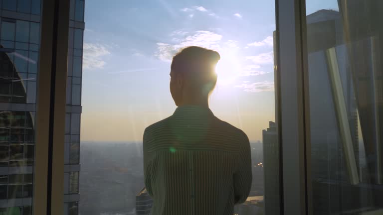 Pensive woman looking at cityscape through window of skyscraper - back view