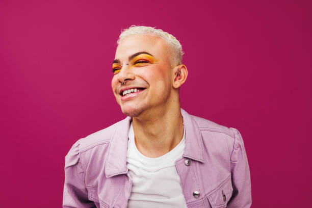 Queer man smiling cheerfully in a studio Queer man smiling cheerfully while standing in a studio. Non-conforming young man wearing colourful eye shadow against a purple background. Happy young man feeling confident in his style. gay man stock pictures, royalty-free photos & images