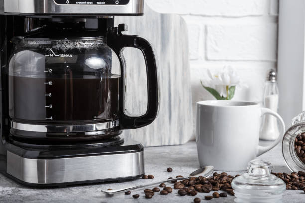 una cafetera eléctrica de goteo negro con una tetera de vidrio prepara una bebida por la mañana. electrodomésticos, una taza blanca y un frasco de frijoles en la mesa de la cocina en la encimera de la casa - coffee black coffee cup coffee bean fotografías e imágenes de stock