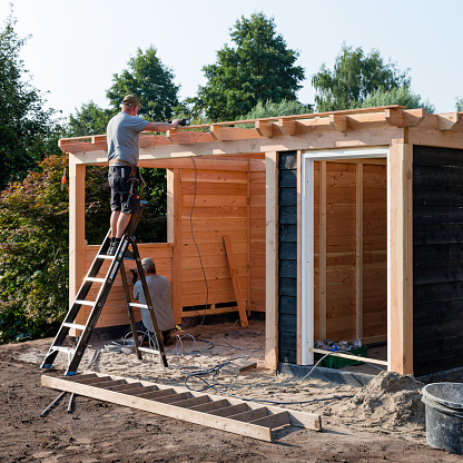 Wierden, Twente, Overijssel, Netherlands, july 26th 2018,  two men building a domestic wooden garden shed in a back yard on a sunny summer day, one is  standing on a ladder mounting timber on the roof, the other is working on the ground, making preparations for electricity - having a garden shed in the back yard is becoming more and more common in the Dutch suburbs