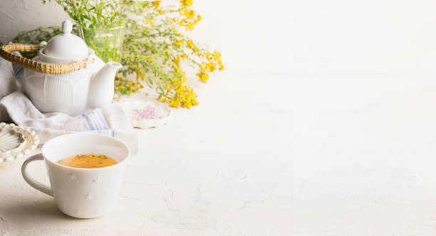 Herbal tea background with cup with yellow tea , tea pot and fresh herbs and flowers on white table at wall. stock photo