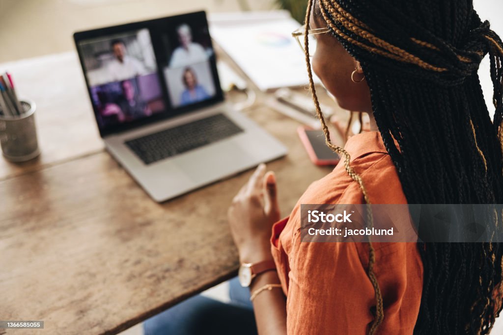 Graphic designer attending a virtual meeting at her desk Graphic designer attending a virtual meeting with her clients. Creative female freelancer using a laptop for a video call.  Young woman working on a new project in her home office. Working At Home Stock Photo