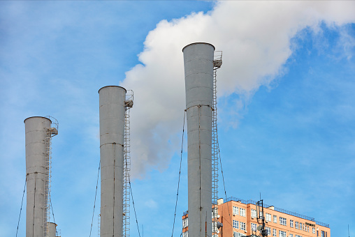 The high three chimneys of the thermal power plant emit steam into the atmosphere against the backdrop of the top of a residential building and a blue sky. Copy space.