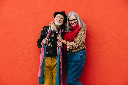 Carefree senior couple laughing while standing together against a red background. Cheerful elderly couple wearing colourful casual clothing. Happy mature couple enjoying life after retirement.