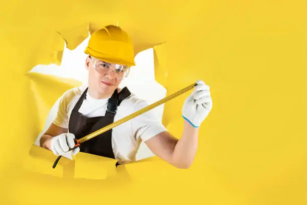 Confident mechanic, construction man. Happy carpenter. Handyman is measuring size of torn hole in yellow background. Senior builder engineer is holding tape-line for measure. Engineering and repair.