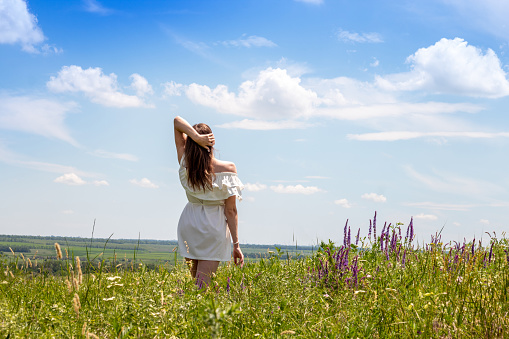 Young woman in white dress stands in the middle of green meadow. Enjoying nature and free life. Selective focus.