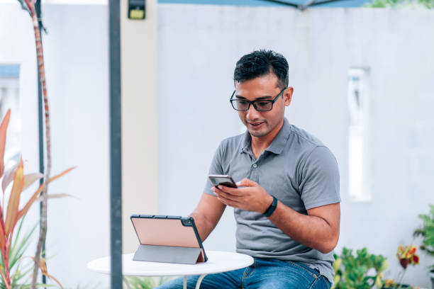 istock Mid adult Muslim man checking emails and text messaging on smart phone while working with digital tablet comfortably in houseplants garden at home. 1366596129