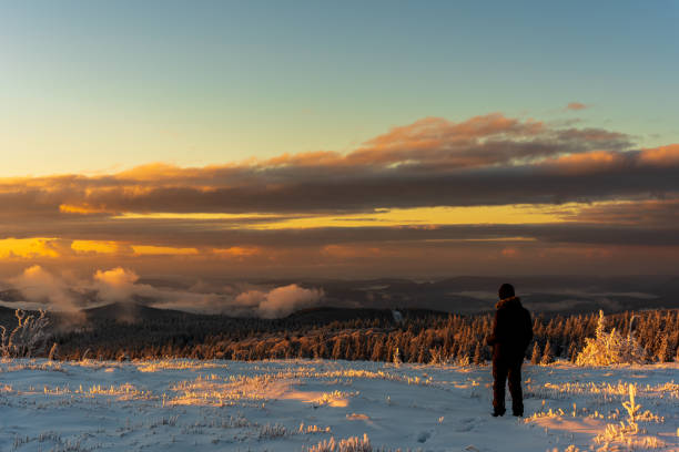 Man standing in snow covered hill landscape overlooking beautiful forest landscape at sunset