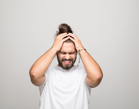 Portrait of bearded young man with hair bun wearing white t-shirt. Angry male student standing with hands in hair, looking down. Studio shot, grey background.