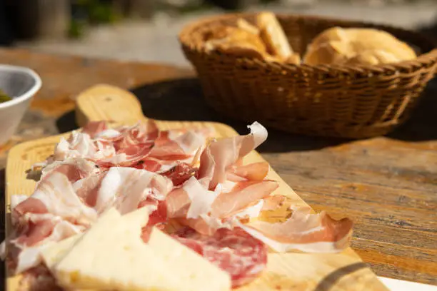 Photo of ham, salami and cheese slices served on a wooden cutting board on a wooden table, blurred vimini bread bowl on the background