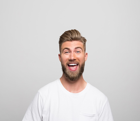 Portrait of handsome bearded young man wearing white t-shirt, smiling at camera. Studio shot, grey background.