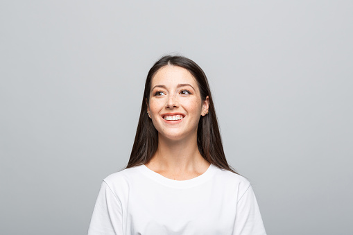 Portrait of beautiful young woman wearing white t-shirt, looking away and smiling. Studio shot, grey background.
