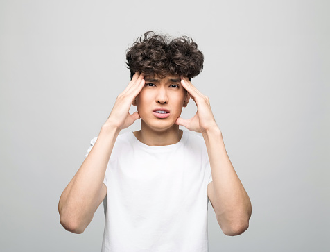 Portrait of sad young man wearing white t-shirt. Male student touching his head. Studio shot, grey background.