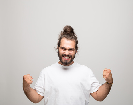 Excited bearded young man with hair bun wearing white t-shirt. Male student standing with raised fists and shouting at camera. Studio shot, grey background.