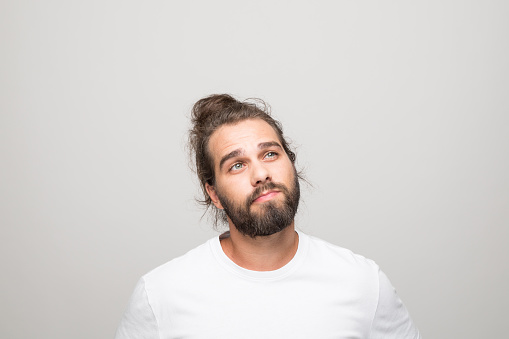 Headshot of bearded young man with hair bun wearing white t-shirt. Thoughtful male student looking away. Studio shot, grey background.