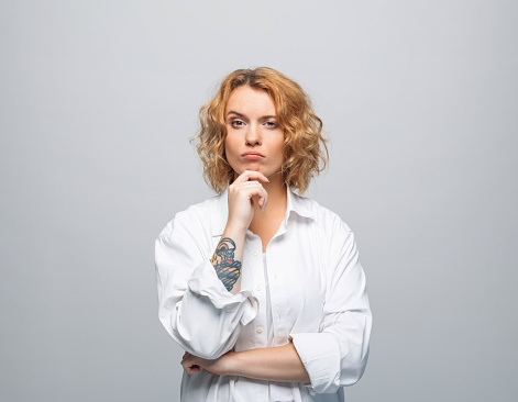 Portrait of beautiful young woman with red curly hair wearing white shirt. Confused female student looking away. Studio shot, grey background.