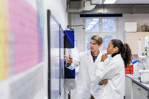 Two researchers in lab coat discussing about chemistry on a whiteboard. Male and female scientists working in a lab looking at formula written on whiteboard.