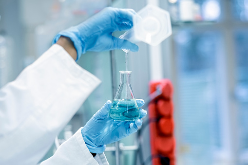 Close-up of a lab technician pouring solution from test tube into a glass flask in laboratory. Scientist working with chemicals for research in a lab.