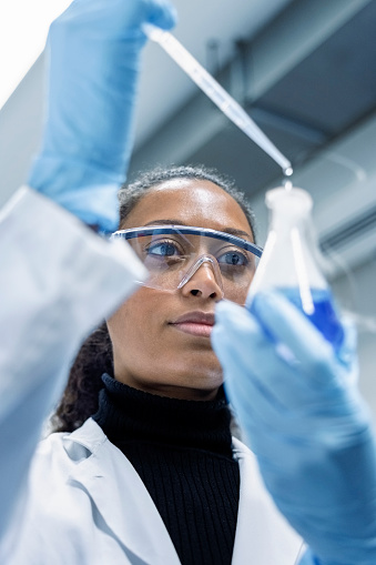 Close-up shot of a woman scientist in lab coat, gloves, and eyeglasses carefully drips chemical from a pipette into glass flask. Lab technician doing experiment in laboratory.