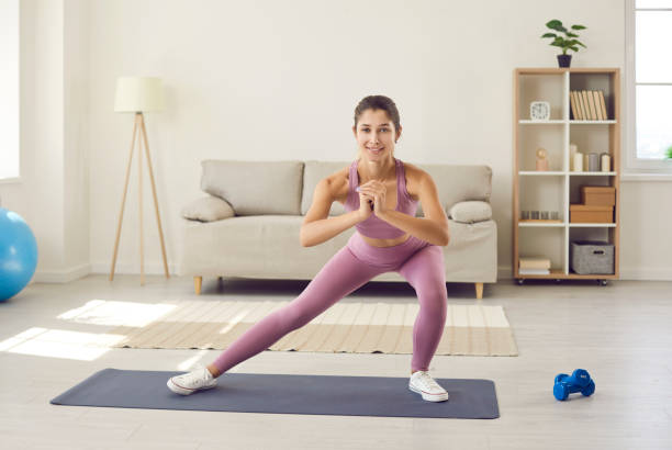 Happy young woman in activewear doing lateral lunges during fitness workout at home Happy beautiful slim woman enjoying routine fitness workout at home. Active young Caucasian lady in modern sports leggings standing on gym mat in living room, smiling and doing side lunge exercise lunge stock pictures, royalty-free photos & images