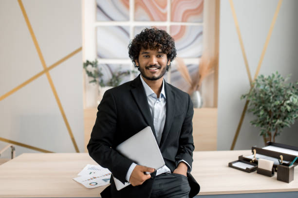 Portrait of a confident successful Indian businessman, manager, IT specialist, stock trader, in formal suit, stands near his desk in the office holds a laptop,looks at the camera with a friendly smile stock photo