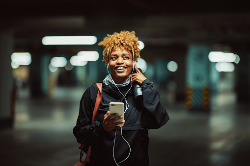 Smiling black woman is standing in a garage and listening to music. She is wearing workout clothes: jacket, headband, scarf and a gym bag. It's evening or night. In one hand the woman is holding a plastic water bottle and in the other hand she is holding a phone connected to the earphones that she is wearing. The garage seems to be empty.