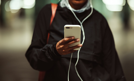 Anonymous serious black woman is standing outside and either typing a text on her mobile phone or choosing music for her training session. She is wearing workout clothes: jacket, headband, scarf and a gym bag. It's evening or night. In one hand the woman is holding a mobile phone connected to the earphones that she is wearing. The garage seems to be empty.