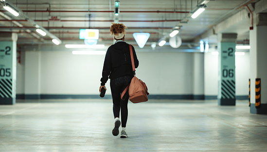 Back view of a black woman who is walking in a garage. She is wearing workout clothes: jacket, headband, scarf, leggings, white socks, sneakers and a gym bag. It's evening or night. In one hand the woman is holding a plastic water bottle. The garage seems to be empty.