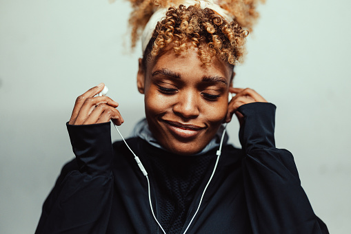 Smiling black woman is standing in front of a wall. She is wearing workout clothes. It's evening or night.  She might be doing a warm-up before training and listening to music because she is wearing earphones. Her eyes are closed.