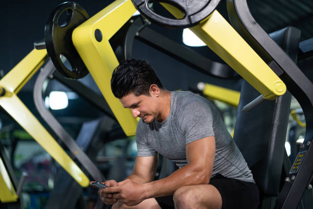 istock Young Caucasian men sitting on weight lifting machine while using smartphone inside of fitness gym. Using smartphone or social network while exercise or workout. 1366610278