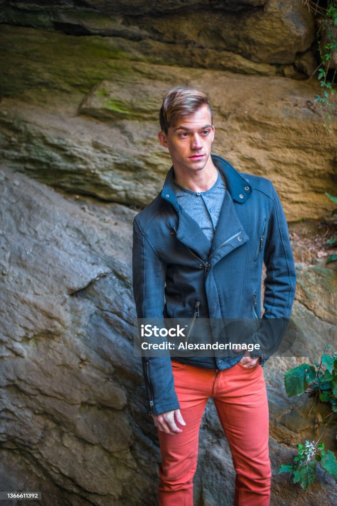 a young man is standing by rocks, relaxing, thinking."r Dressing in a blue slim fit jacket,  red jeans, a young man is standing by rocks, relaxing, thinking."r Brown Stock Photo