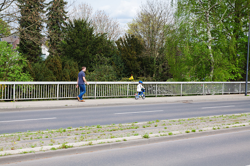 Man and a child on bicycle are crossing Mendener Bruecke in Muelheim Ruhr in spring season.  Man is walking behind cycling boy, Boy is wearing a football shirt In background are trees at river Ruhr
