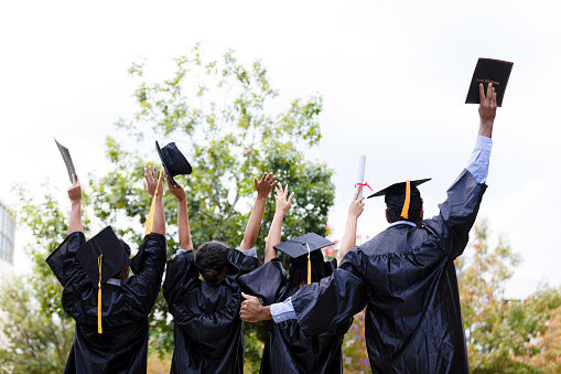 A rear view photograph of four graduates in their caps and gowns.  The hold caps and diplomas in their upraised hands.