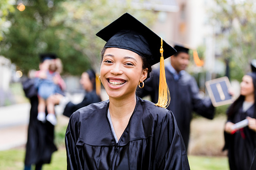 While her friends talk in the background after the graduation ceremony, the young adult woman smiles for a photo.
