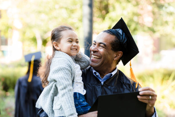 Little girl makes graduating grandfather laugh with silly face The little preschool age girl makes a silly face. Her mature adult grandfather smiles and laughs. He has just graduated. graduation man stock pictures, royalty-free photos & images