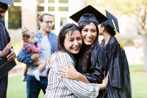 A mid adult mother and her young adult graduating daughter pose cheek to cheek with big smiles for a photo taken by an unseen person.
