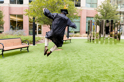Rear view of a man excitedly jumping, celebrating his recent graduation from college. The man is jumping on a lawn outside of a university building.