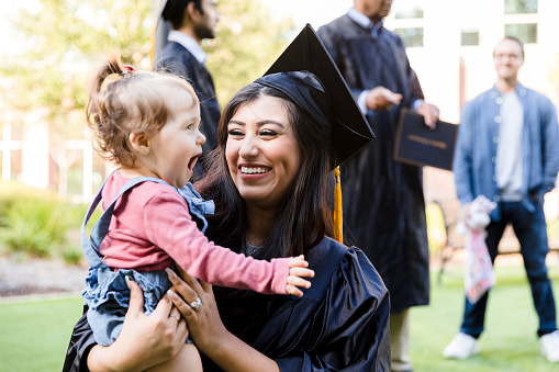 The young adult mother, in her cap and gown, smiles at her cute baby girl's silliness.