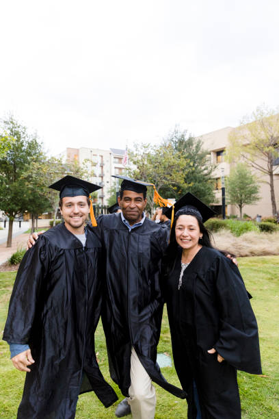 A group of five multiracial high school graduates standing together outside the school, wearing their caps and gowns, smiling and waving to the camera.