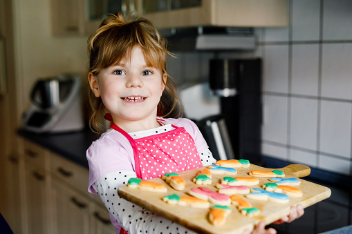 Cute little toddler girl holding fresh baked homemade Easter or spring cookies at home indoors. Adorable blond child with apron with bunny and carrot cookie in domestic kitchen