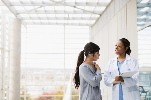 The young adult female patient stops the mid adult female doctor in the hospital walkway to ask about a pain in her shoulder.