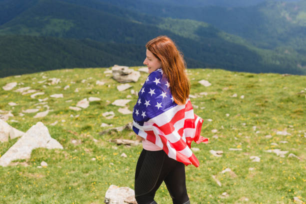 girl with american flag looking out at landscape. young woman stands in the mountains, holding the us flag in her arms high. flag fluttering in the wind - ridge mountain wilderness area poland imagens e fotografias de stock