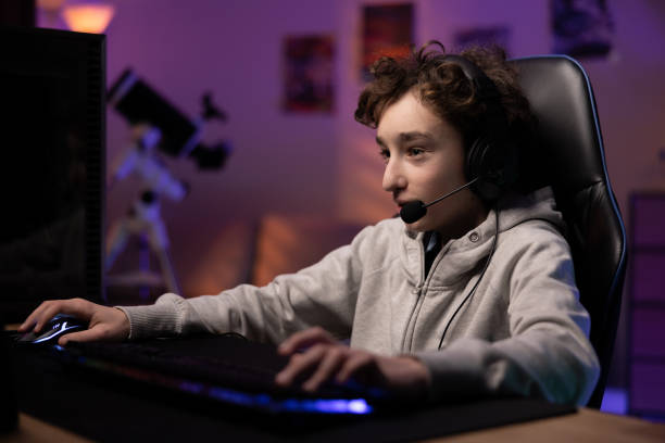 istock Focused young boy with curly hair spends time at computer playing multiplayer video games with team, talks to people through headset, dark room night lit by neon led lights 1366633395