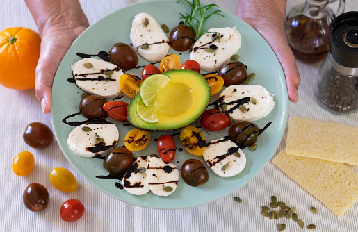 Top view of caprese salad. Female hands hold a plate with cheese mozzarella and small tomatoes, pepper and balsamic. Half avocado to complete a healthy meal