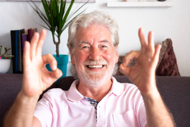 istock Cheerful white-haired senior man sitting at home on sofa making ok sign with hands, looking smiling into camera. Positive message from an elderly youthful grandfather 1366635014